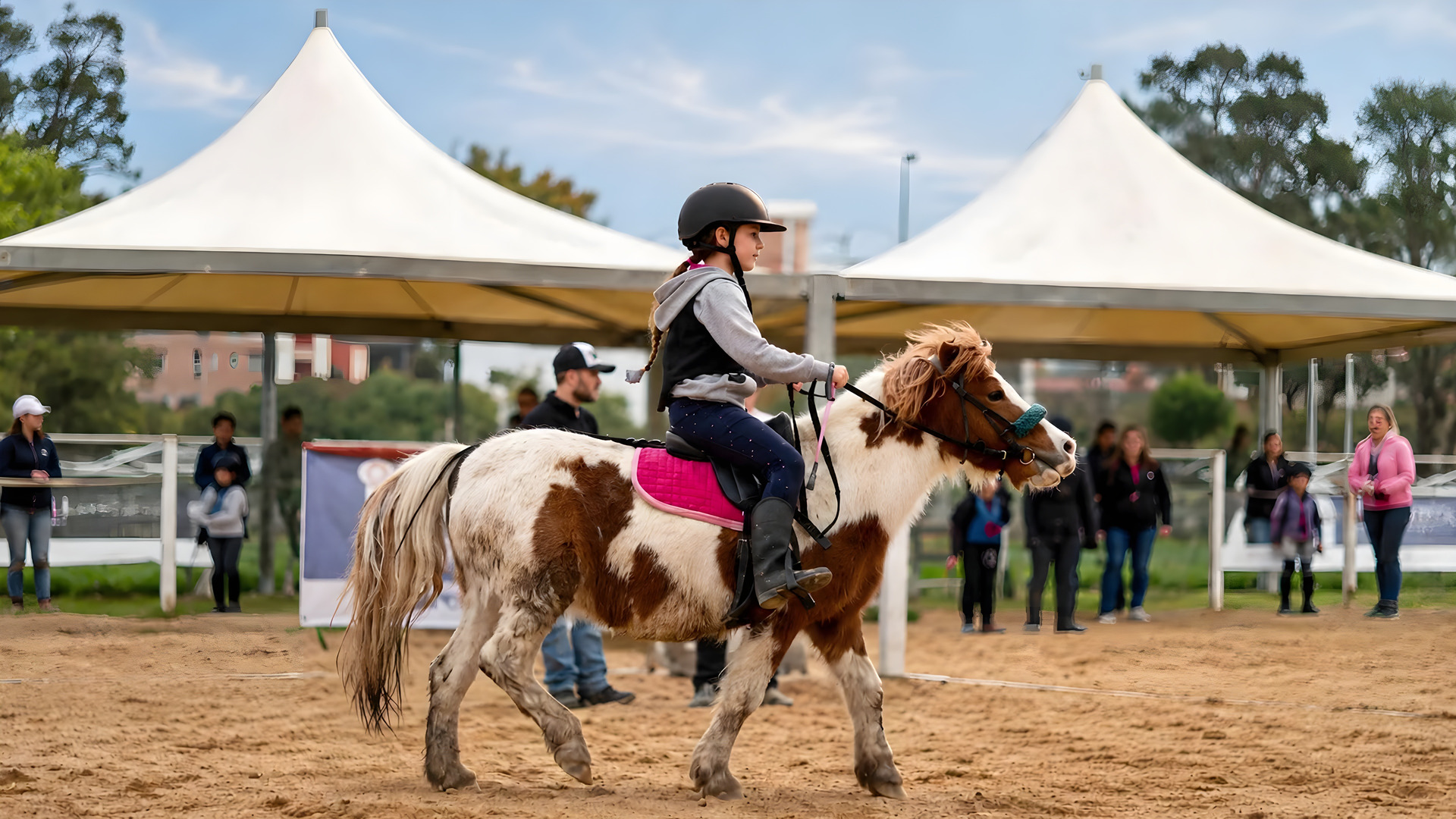 Una bambina attraversa al passo il campo in sabbia su un pony pezzato, con i monti alle spalle